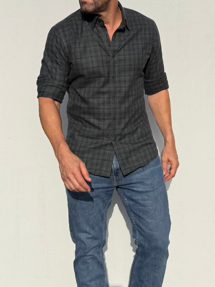 A man wearing a dark gray and black plaid button-down shirt with rolled-up sleeves and blue jeans, standing against a plain off-white wall.