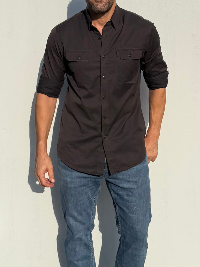 A man wearing a dark brown long-sleeve button-down shirt with chest flap pockets and blue jeans, standing against a light gray wall.